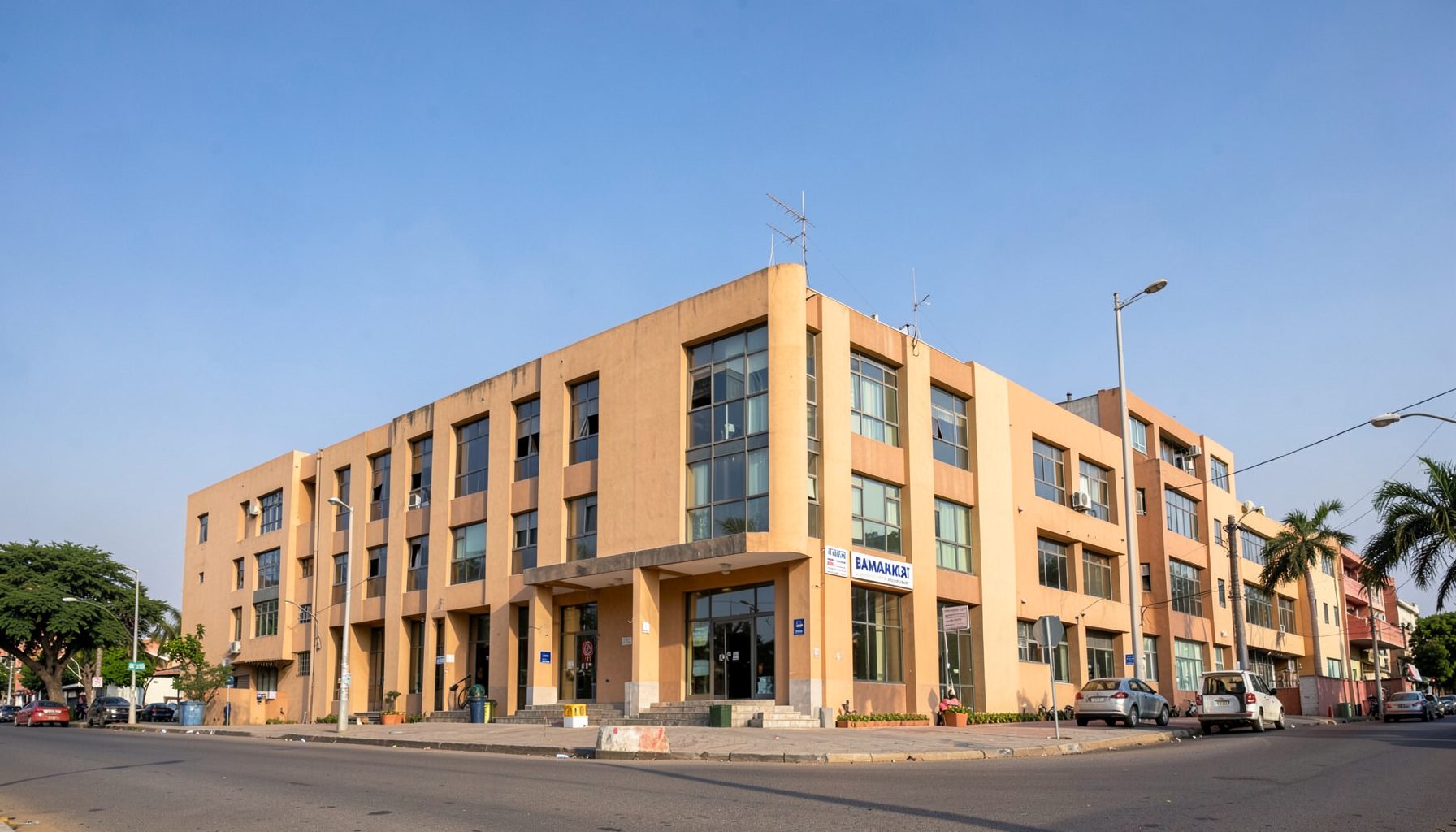 Широка панорама на административна зграда во Бамако со топли земјени тонови, песочна фасада и сино небо, пренесува институционална атмосфера